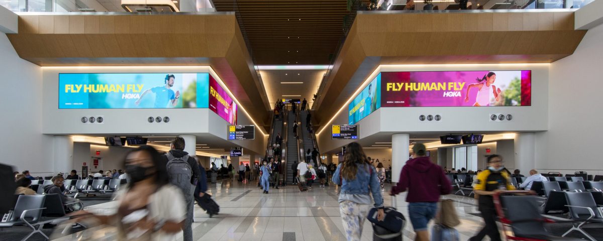 Busy airport terminal with travelers walking past large digital advertising screens displaying the “Fly Human Fly” HOKA running campaign near escalators.