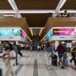 Busy airport terminal with travelers walking past large digital advertising screens displaying the “Fly Human Fly” HOKA running campaign near escalators.