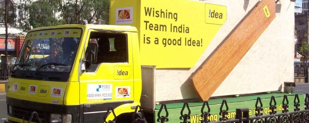 A branded outdoor mobile van advertising campaign by Idea features a bright yellow promotional vehicle with large billboard-style panels and bold messaging supporting Team India. The mobile advertising truck is designed for high visibility roadshows, outdoor brand promotions, and citywide marketing activations targeting mass public engagement.