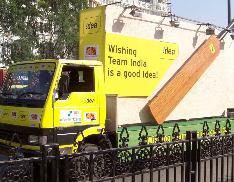 A branded outdoor mobile van advertising campaign by Idea features a bright yellow promotional vehicle with large billboard-style panels and bold messaging supporting Team India. The mobile advertising truck is designed for high visibility roadshows, outdoor brand promotions, and citywide marketing activations targeting mass public engagement.