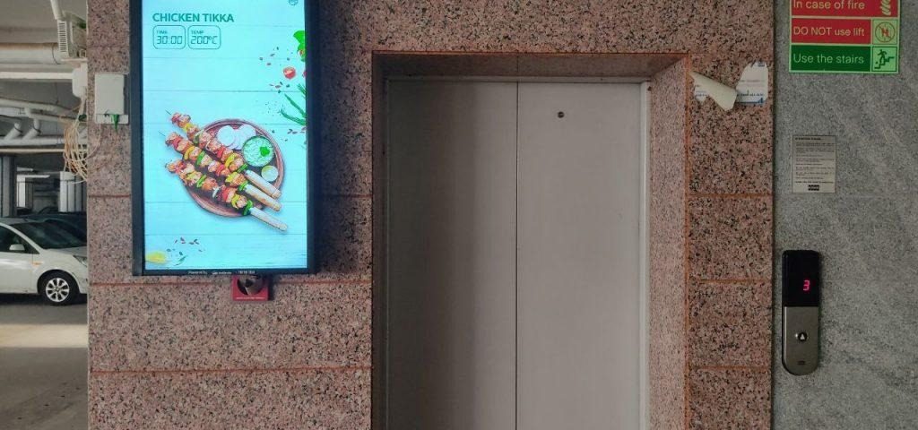 An elevator branding campaign features a digital advertising screen installed beside an elevator in a commercial parking area, displaying a food promotion with vibrant visuals and pricing information. The indoor advertising setup uses elevator branding and digital display marketing to capture audience attention in high footfall residential and commercial spaces.