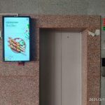 An elevator branding campaign features a digital advertising screen installed beside an elevator in a commercial parking area, displaying a food promotion with vibrant visuals and pricing information. The indoor advertising setup uses elevator branding and digital display marketing to capture audience attention in high footfall residential and commercial spaces.