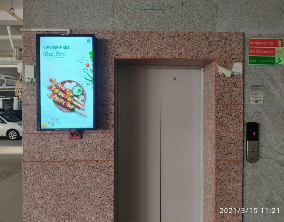 An elevator branding campaign features a digital advertising screen installed beside an elevator in a commercial parking area, displaying a food promotion with vibrant visuals and pricing information. The indoor advertising setup uses elevator branding and digital display marketing to capture audience attention in high footfall residential and commercial spaces.