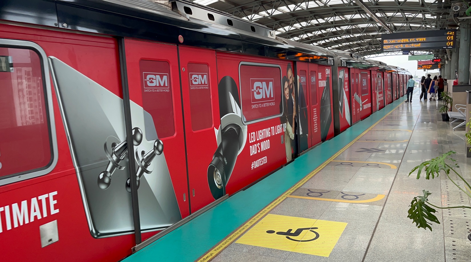 Red metro train fully wrapped with GM LED lighting advertisement at an elevated metro station platform.