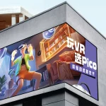 Corner-mounted digital billboard on a modern building displaying an immersive VR gaming advertisement featuring a person using a virtual reality headset.