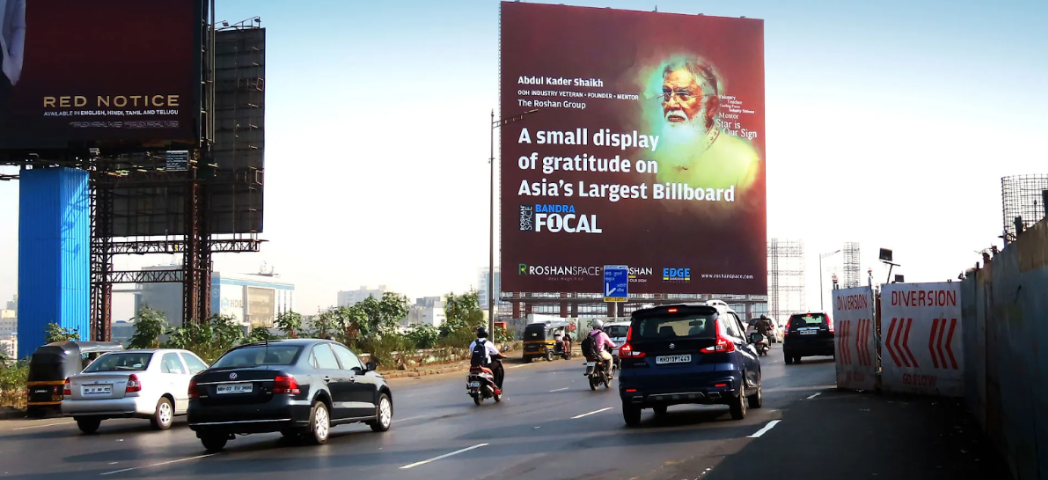 Large roadside billboard with a portrait and text, viewed from a busy multi-lane road with cars and motorcycles