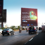 Large roadside billboard with a portrait and text, viewed from a busy multi-lane road with cars and motorcycles
