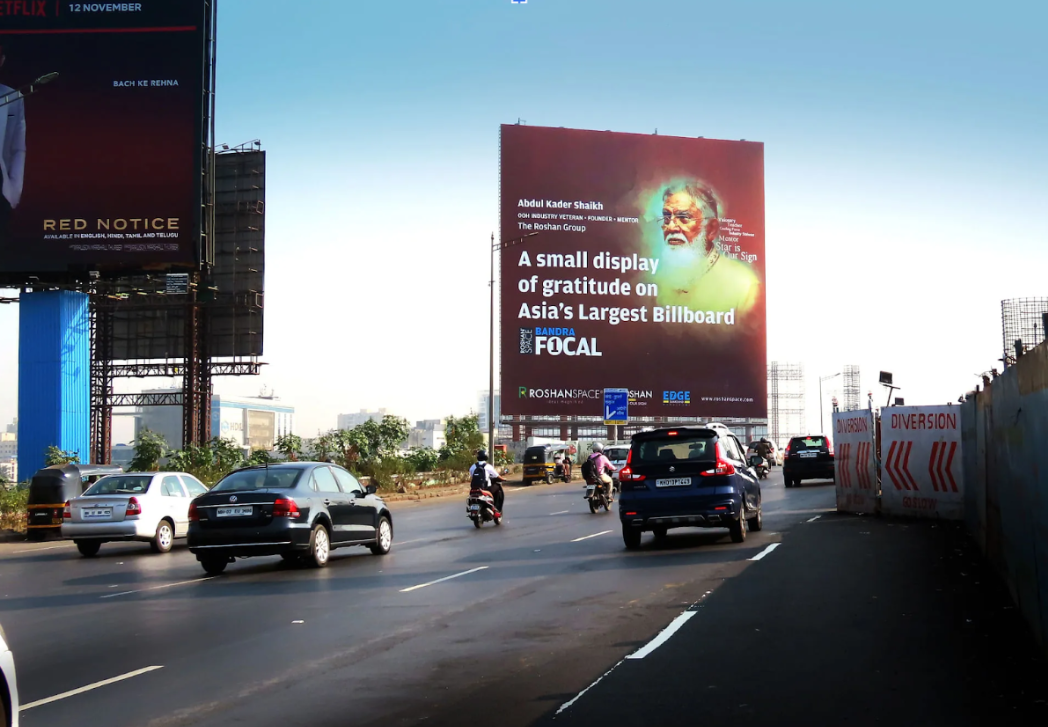 Large roadside billboard with a portrait and text, viewed from a busy multi-lane road with cars and motorcycles