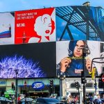 Large digital billboards displaying multiple brand advertisements at a busy city intersection with traffic and pedestrians visible