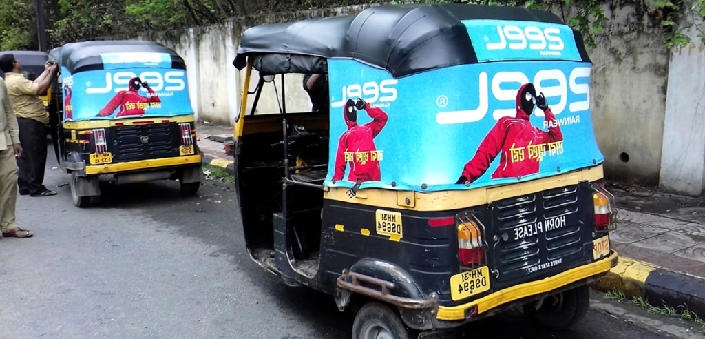 Auto rickshaws parked on a city street with rear-panel advertisement wraps displaying a fashion campaign in bright blue and red colors.