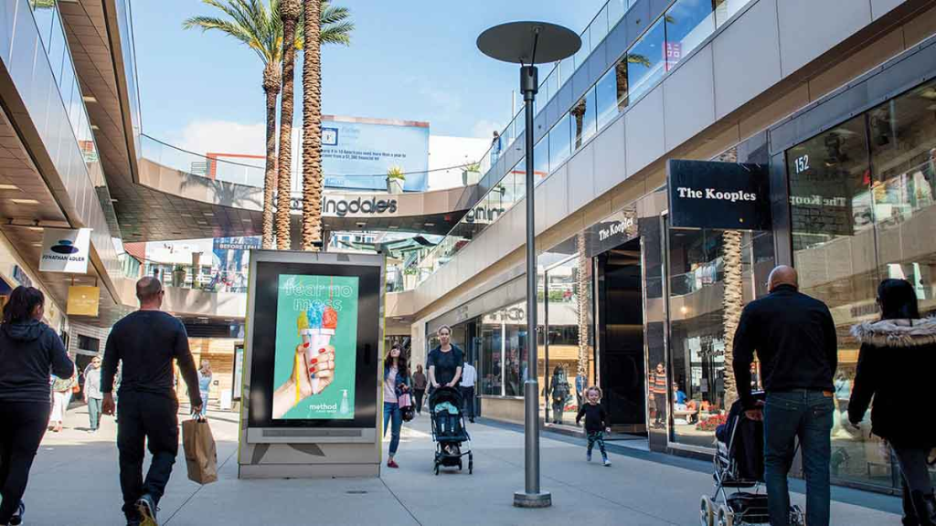 Outdoor shopping mall walkway featuring a freestanding digital advertising kiosk displaying a colorful beverage campaign among pedestrians.