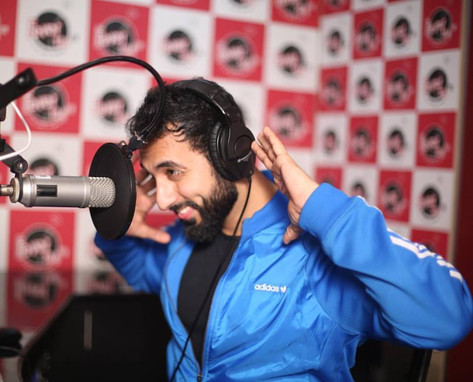 Radio host wearing headphones speaking into a microphone inside a recording studio with branded backdrop.