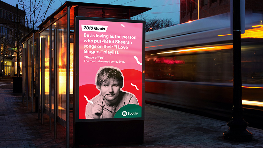 Illuminated bus shelter advertisement featuring Spotify’s 2018 Goals campaign highlighting Ed Sheeran’s “Shape of You,” displayed on a digital panel at night with a passing bus in motion blur.