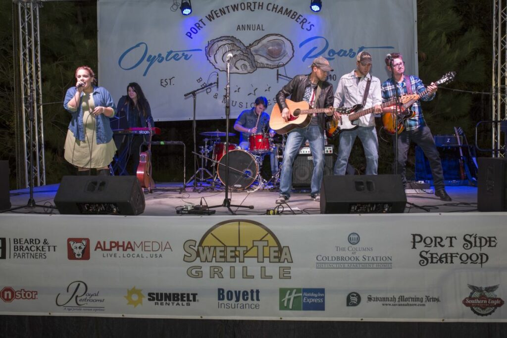 Live band performing on stage at an outdoor event with sponsor logos displayed on banners across the stage front.