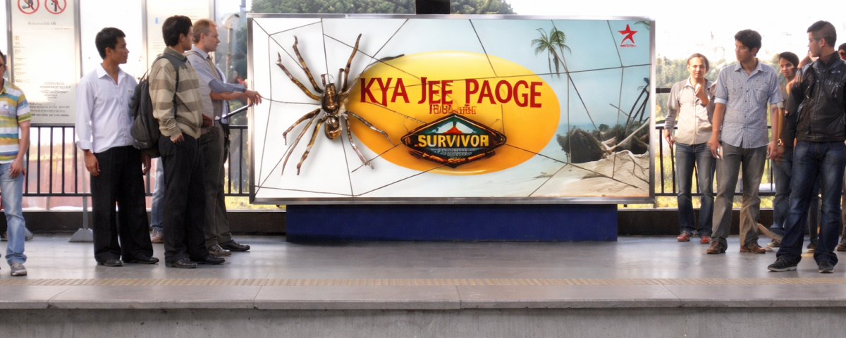 People standing on a metro platform in front of a large billboard with a spider graphic and “Kya Jee Paoge” text