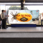 People standing on a metro platform in front of a large billboard with a spider graphic and “Kya Jee Paoge” text
