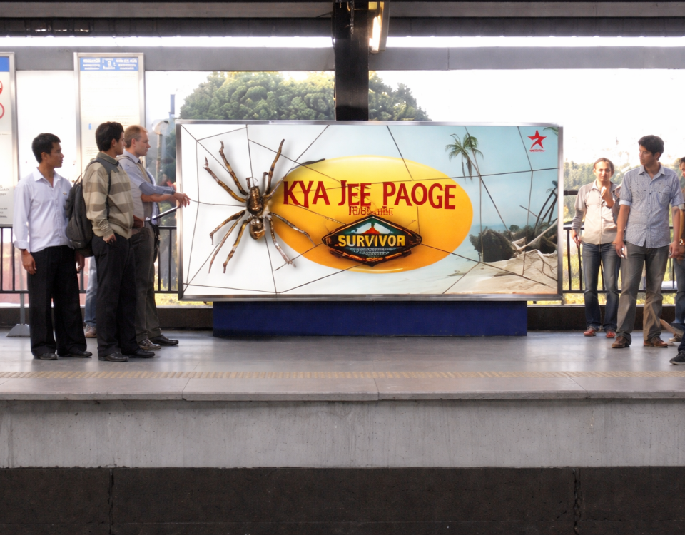 People standing on a metro platform in front of a large billboard with a spider graphic and “Kya Jee Paoge” text