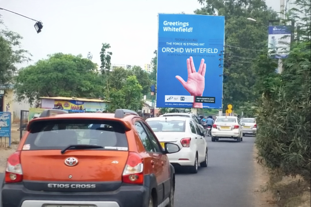 Large roadside billboard in Whitefield displaying a blue advertisement with raised hand graphic and greeting message