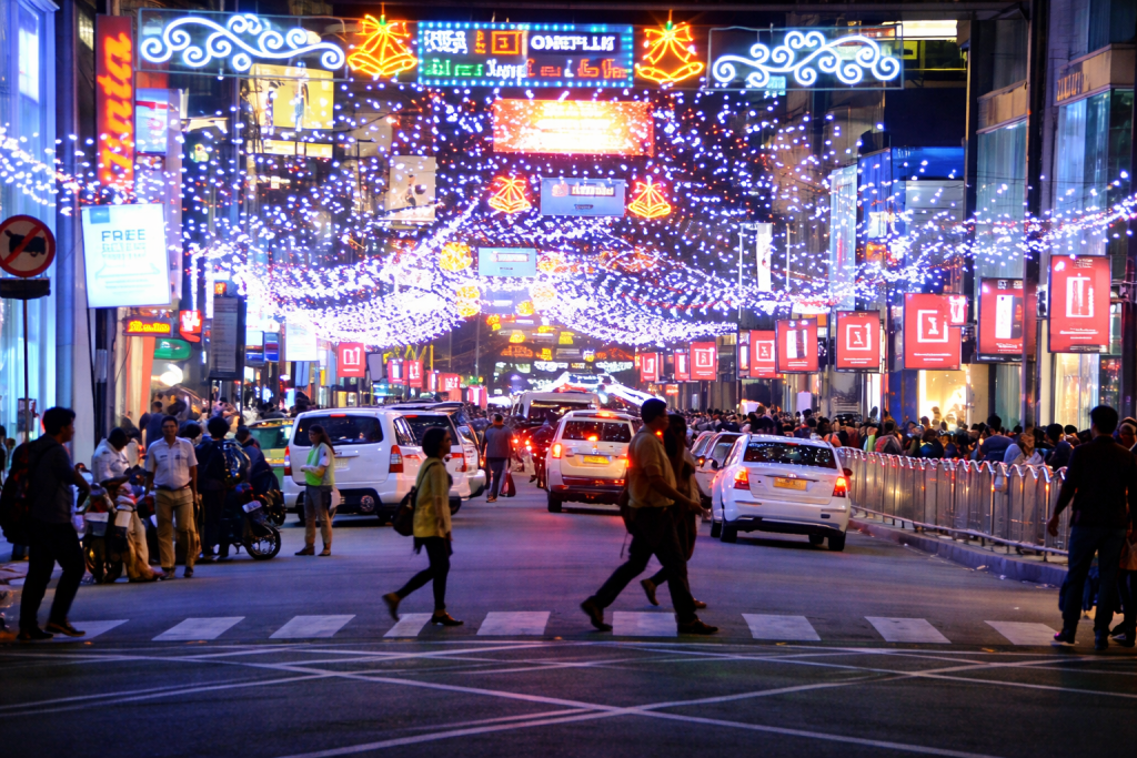 Busy city street at night decorated with festive lights and illuminated signage, with cars and pedestrians moving through a crowded shopping district.