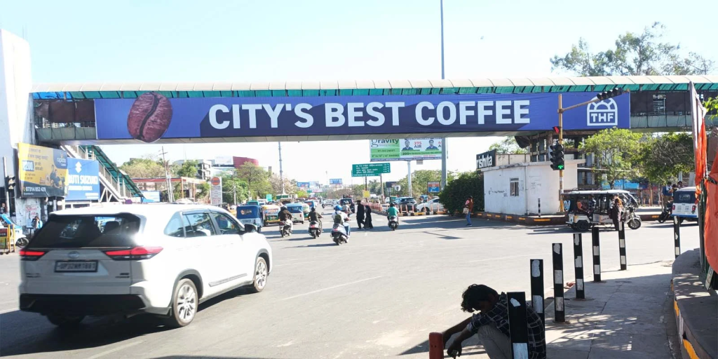 Pedestrian bridge over busy road displaying “City’s Best Coffee” banner advertisement with traffic moving below.