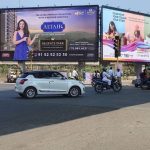 Large dual-sided billboards at a busy urban intersection featuring real estate and lifestyle advertisements, with cars, auto-rickshaws, and motorbikes navigating the junction.