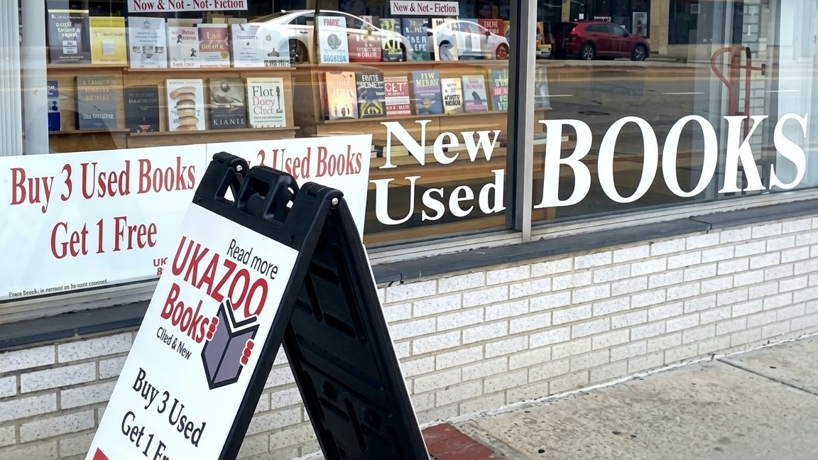 Bookstore window displaying “New Used Books” signage with a sidewalk stand promoting buy three get one free offer.