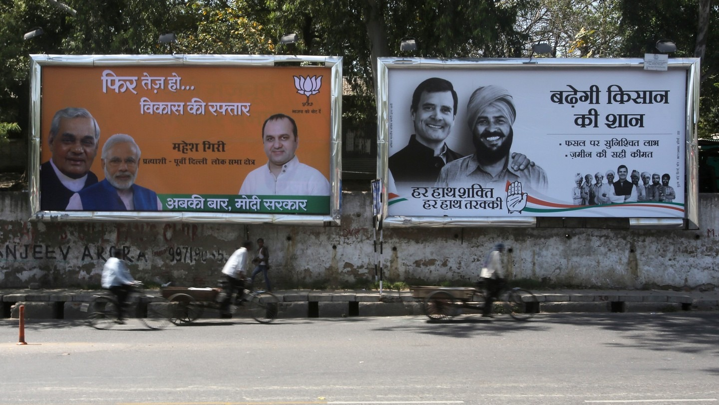 BJP and Congress election campaign billboards in Hindi displayed side by side on a Delhi street, with cyclists passing in the foreground
