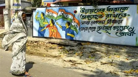 Woman in saree walking past a Trinamool Congress (TMC) election campaign mural with Bengali text painted on a wall in West Bengal, India