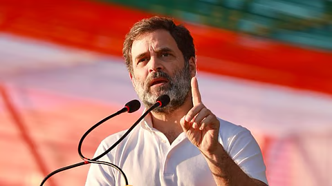 Indian politician speaking at a public rally with microphones, raising one finger, against a red background