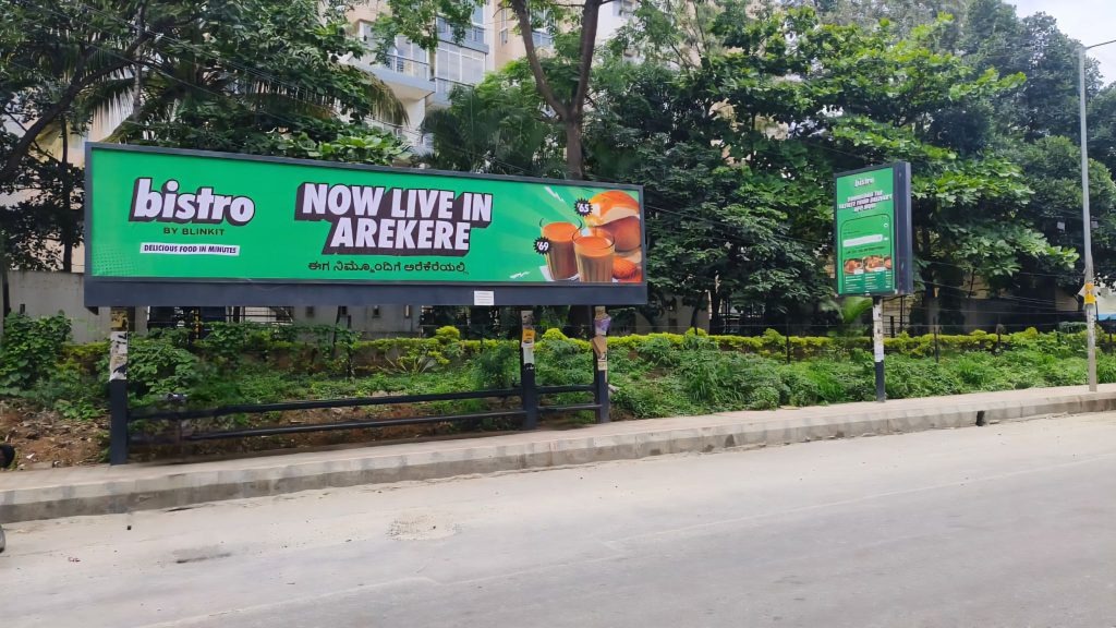 Green roadside billboard promoting Bistro by Blinkit announcing “Now Live in Arekere” along a tree-lined urban street.