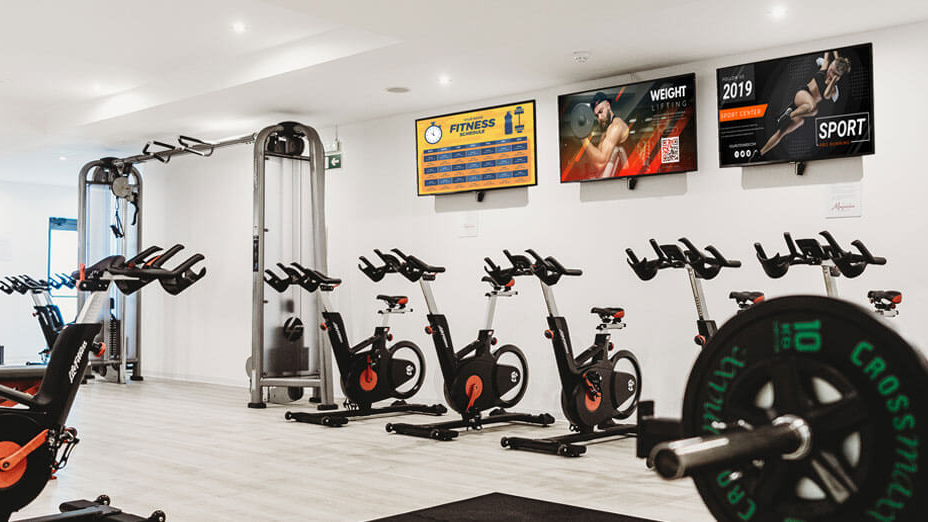 Gym Interior Digital Screens Promoting Fitness Content Digital advertising screens mounted on a gym wall displaying fitness promotions above stationary bikes and workout equipment.
