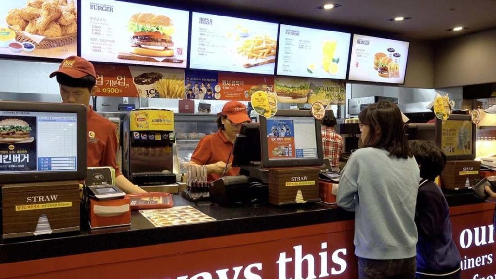 Customers ordering food at a busy fast-food restaurant counter with digital menu boards displaying burgers, fries, and meal options.