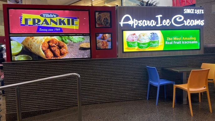 Bright digital advertising displays for Tibbs Frankie and Apsara Ice Creams at a food court seating area.
