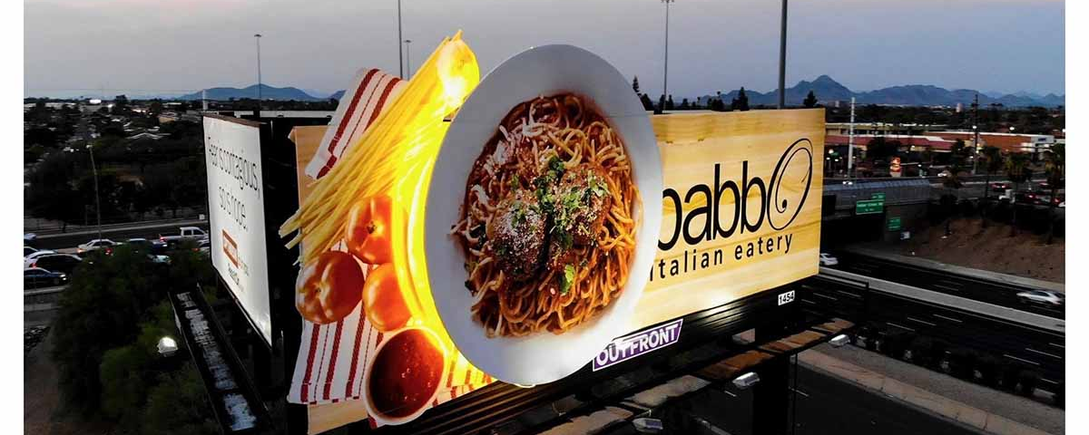 3D highway billboard advertising an Italian restaurant featuring an oversized plate of spaghetti with meatballs and illuminated pasta elements extending beyond the billboard.