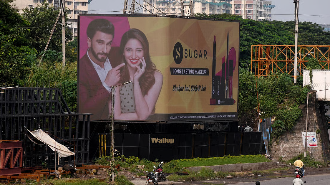 SUGAR Cosmetics billboard showcasing beauty products and brand ambassadors mounted on a roadside structure surrounded by greenery and urban buildings.