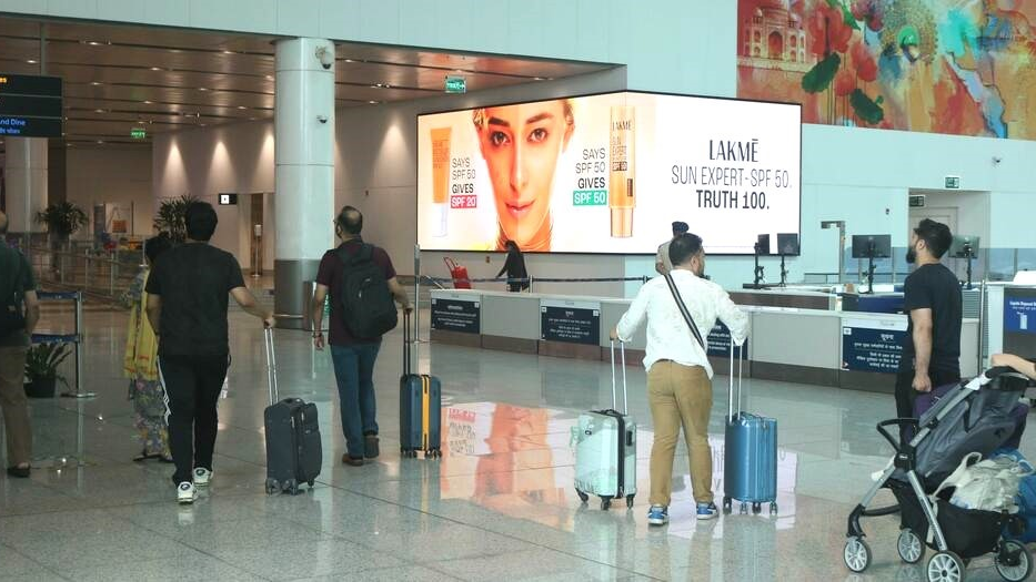 Travelers with luggage walking through an airport terminal past a large Lakmé Sun Expert SPF 50 digital advertisement near the check-in counters.
