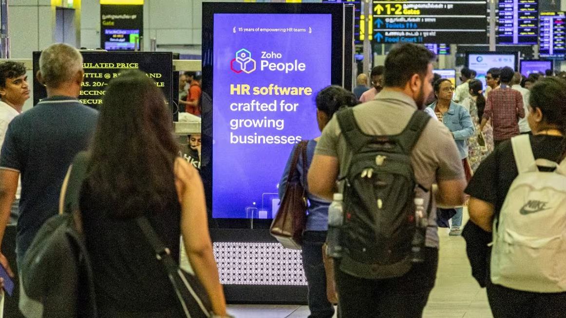Travelers walking through a busy airport terminal past a digital advertising kiosk promoting Zoho People HR software.