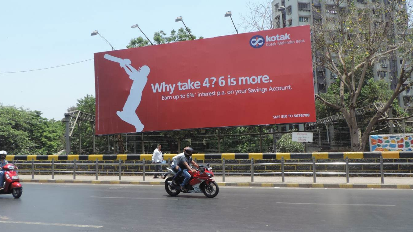 Kotak Mahindra Bank billboard promoting savings account interest rates placed above a city roadway with motorcyclists riding below.