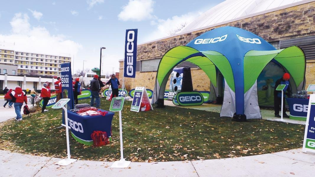 GEICO branded outdoor activation booth with promotional tent, signage, and staff engaging visitors during a marketing event.