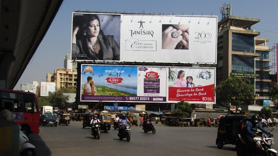 Large Tanishq jewellery billboard mounted above a busy city intersection with multiple vehicles, auto-rickshaws, and surrounding commercial buildings.