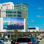 A large roadside billboard mounted on the side of a multi-story hospital building displays a cruise advertisement reading “Our Biggest Offer Is Here,” overlooking a busy highway with moving cars under a clear blue sky.
