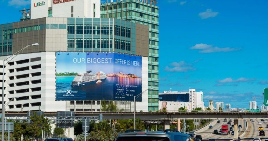 A large roadside billboard mounted on the side of a multi-story hospital building displays a cruise advertisement reading “Our Biggest Offer Is Here,” overlooking a busy highway with moving cars under a clear blue sky.