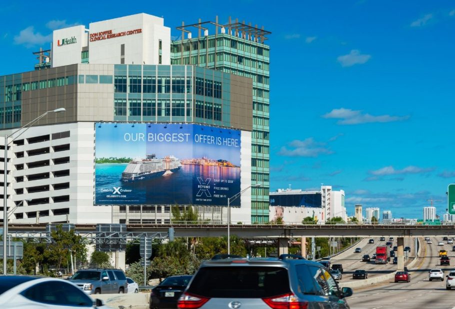 A large roadside billboard mounted on the side of a multi-story hospital building displays a cruise advertisement reading “Our Biggest Offer Is Here,” overlooking a busy highway with moving cars under a clear blue sky.