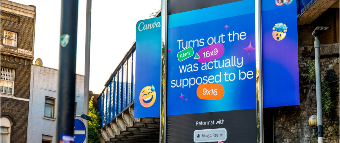 A large outdoor billboard advertisement placed under a bridge in an urban street setting, promoting Canva’s Magic Resize feature. The design uses a bright blue gradient background with playful icons and text explaining how a 16:9 format can be converted into 9:16. The billboard is mounted on a sidewalk near traffic signals, buildings, and a passing red bus, highlighting real-world outdoor advertising placement.