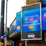 A large outdoor billboard advertisement placed under a bridge in an urban street setting, promoting Canva’s Magic Resize feature. The design uses a bright blue gradient background with playful icons and text explaining how a 16:9 format can be converted into 9:16. The billboard is mounted on a sidewalk near traffic signals, buildings, and a passing red bus, highlighting real-world outdoor advertising placement.
