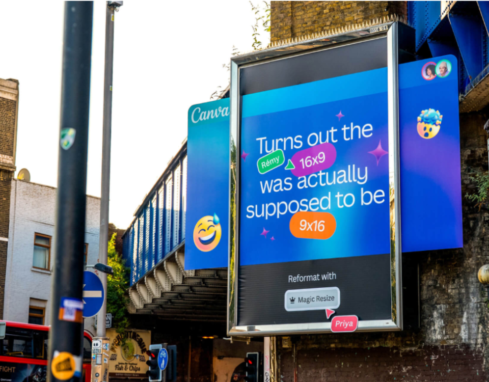 A large outdoor billboard advertisement placed under a bridge in an urban street setting, promoting Canva’s Magic Resize feature. The design uses a bright blue gradient background with playful icons and text explaining how a 16:9 format can be converted into 9:16. The billboard is mounted on a sidewalk near traffic signals, buildings, and a passing red bus, highlighting real-world outdoor advertising placement.