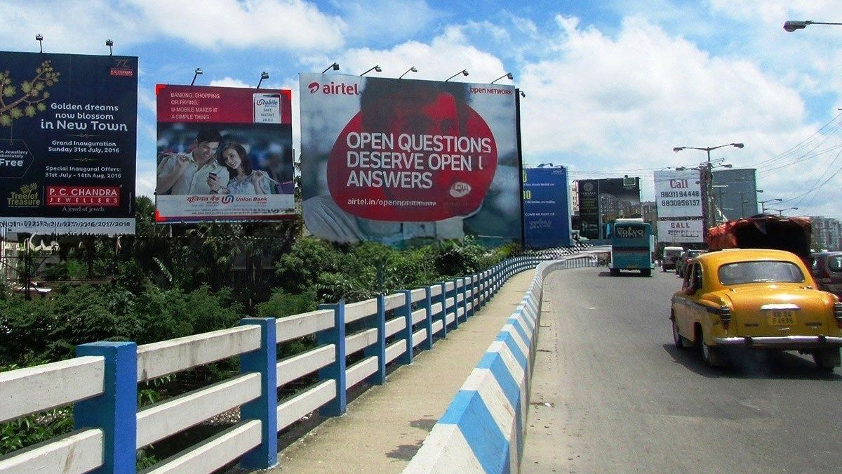 A view from a flyover featuring large Airtel and Union Bank billboards. A classic yellow taxi drives toward the camera under a bright, cloudy sky.