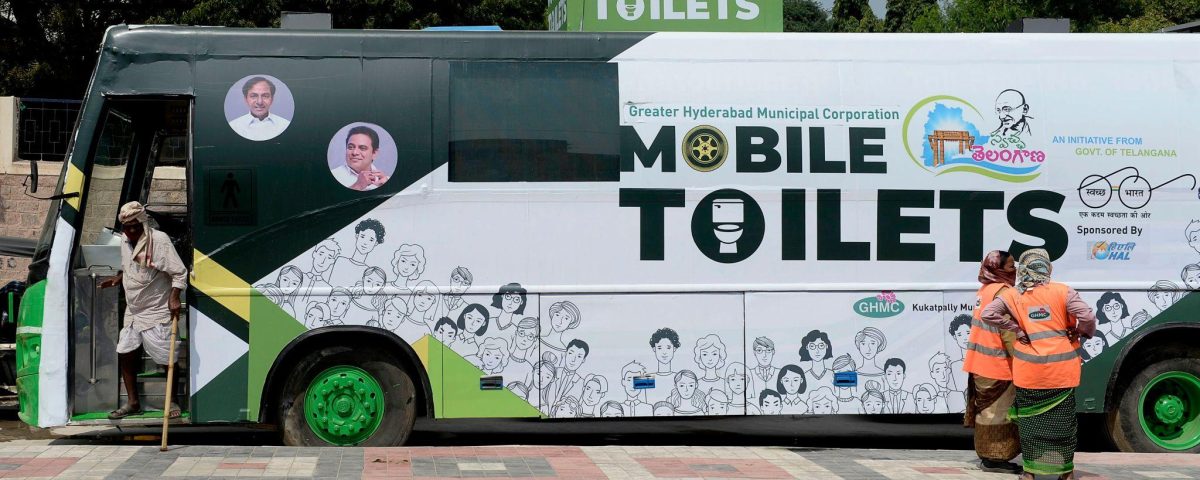 A large green-and-white bus labeled “Mobile Toilets” parked roadside, part of a public sanitation initiative by Greater Hyderabad Municipal Corporation, with illustrations of people on its exterior. A man is stepping into the bus from the left, while two sanitation workers in orange safety vests stand near the rear. Trees and a clear sky form the background.