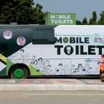 A large green-and-white bus labeled “Mobile Toilets” parked roadside, part of a public sanitation initiative by Greater Hyderabad Municipal Corporation, with illustrations of people on its exterior. A man is stepping into the bus from the left, while two sanitation workers in orange safety vests stand near the rear. Trees and a clear sky form the background.