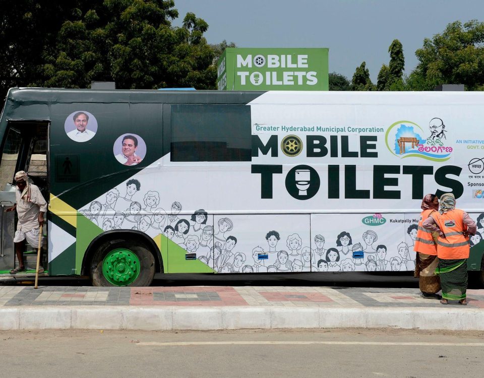 A large green-and-white bus labeled “Mobile Toilets” parked roadside, part of a public sanitation initiative by Greater Hyderabad Municipal Corporation, with illustrations of people on its exterior. A man is stepping into the bus from the left, while two sanitation workers in orange safety vests stand near the rear. Trees and a clear sky form the background.